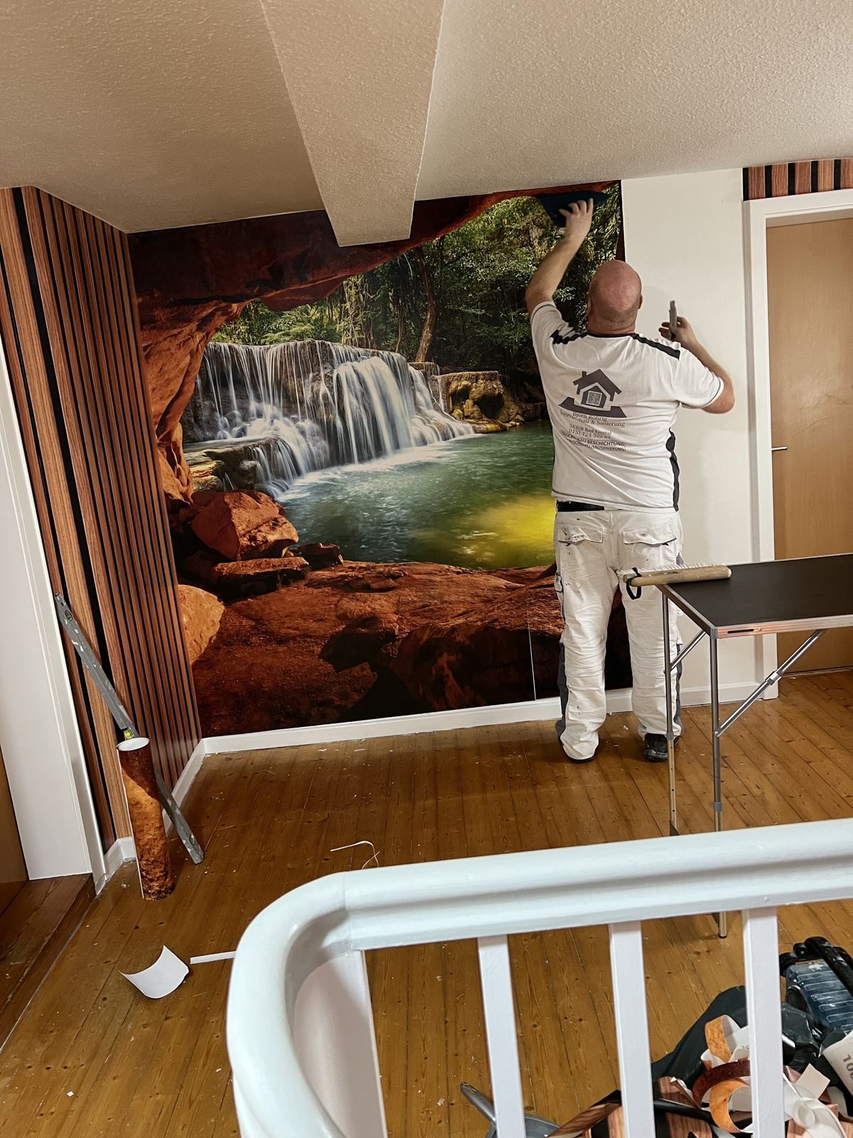 Björn Röhrig bringt eine Fototapete im Treppenhaus an, daneben Holzlamellen
